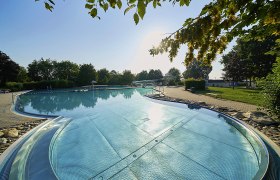 Ein Freibad mit klarem Wasser, umgeben von Bäumen und einem sonnigen Himmel.