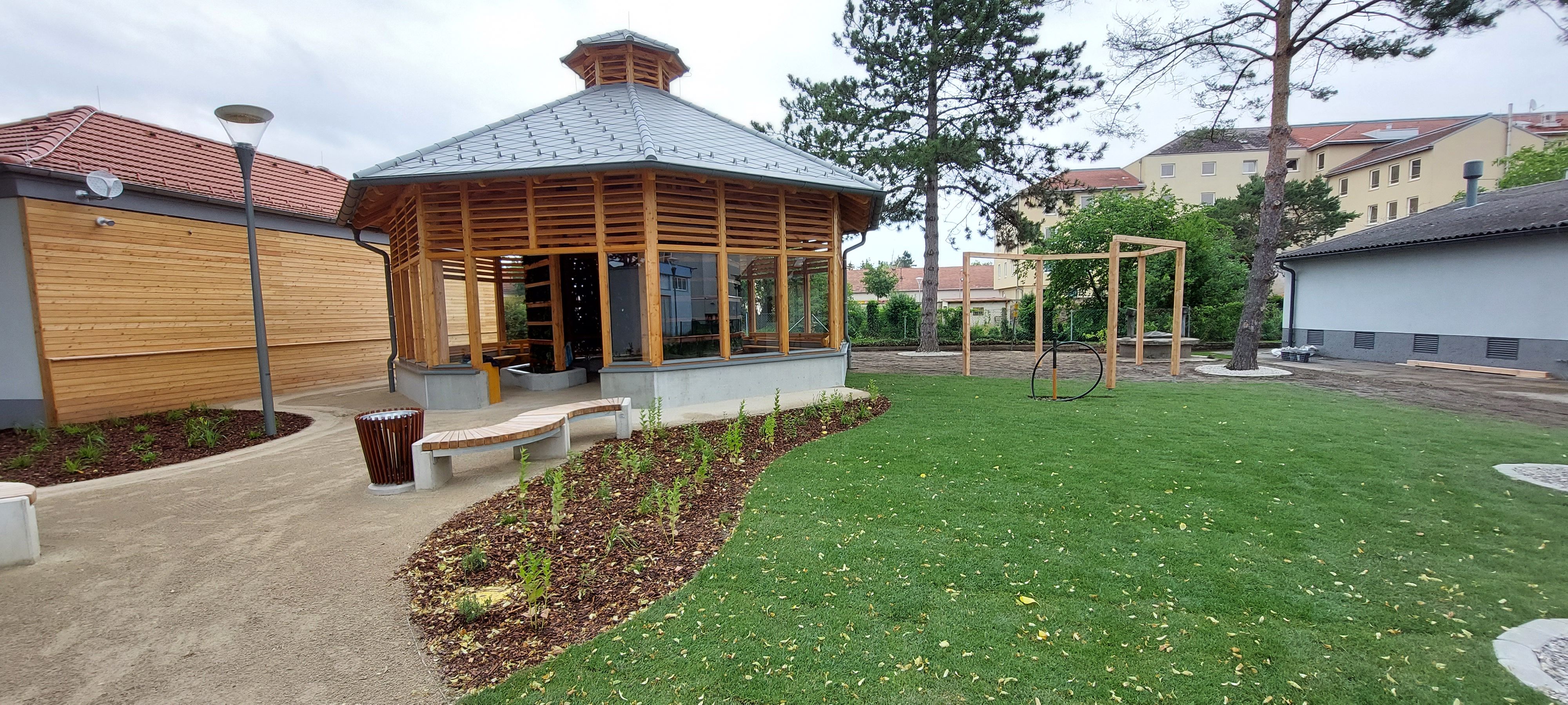 Ein modernes Inhalatorium mit Holzkonstruktion und Glasfenstern in einem gepflegten Garten mit Rasen und Bäumen.