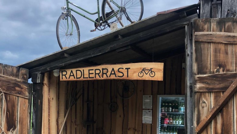 Ein h&ouml;lzerner Unterstand mit einem Fahrrad auf dem Dach und einem K&uuml;hlschrank mit Getr&auml;nken. Ein Schild mit der Aufschrift 'Radlerrast'.