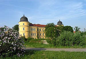 Gelbes Schloss mit zwei T&uuml;rmen und rotem Dach, umgeben von B&auml;umen und Blumen.