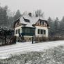 Ein verschneites Haus im traditionellen Stil mit gr&uuml;nen Fensterl&auml;den, umgeben von B&auml;umen und einer schneebedeckten Stra&szlig;e.