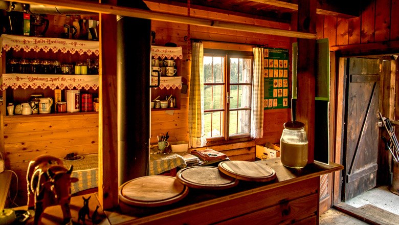 Gem&uuml;tliche Holzh&uuml;tte mit rustikaler Einrichtung, einem Fenster mit Vorh&auml;ngen und einem offenen T&uuml;rbereich.