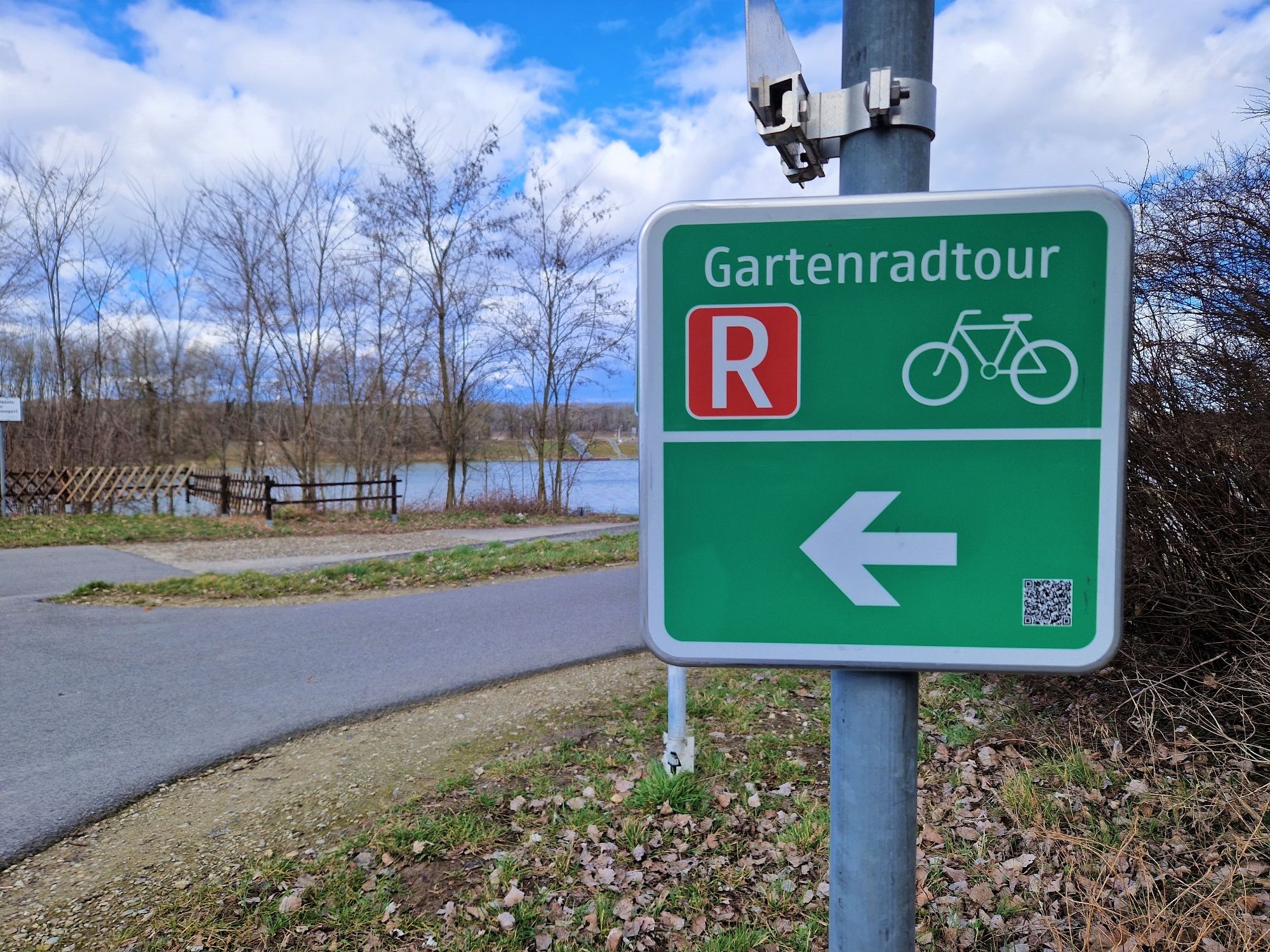 Ein grünes Schild mit der Aufschrift 'Gartenradtour' und einem Fahrradsymbol zeigt nach links. Im Hintergrund ist ein Weg und ein See zu sehen.