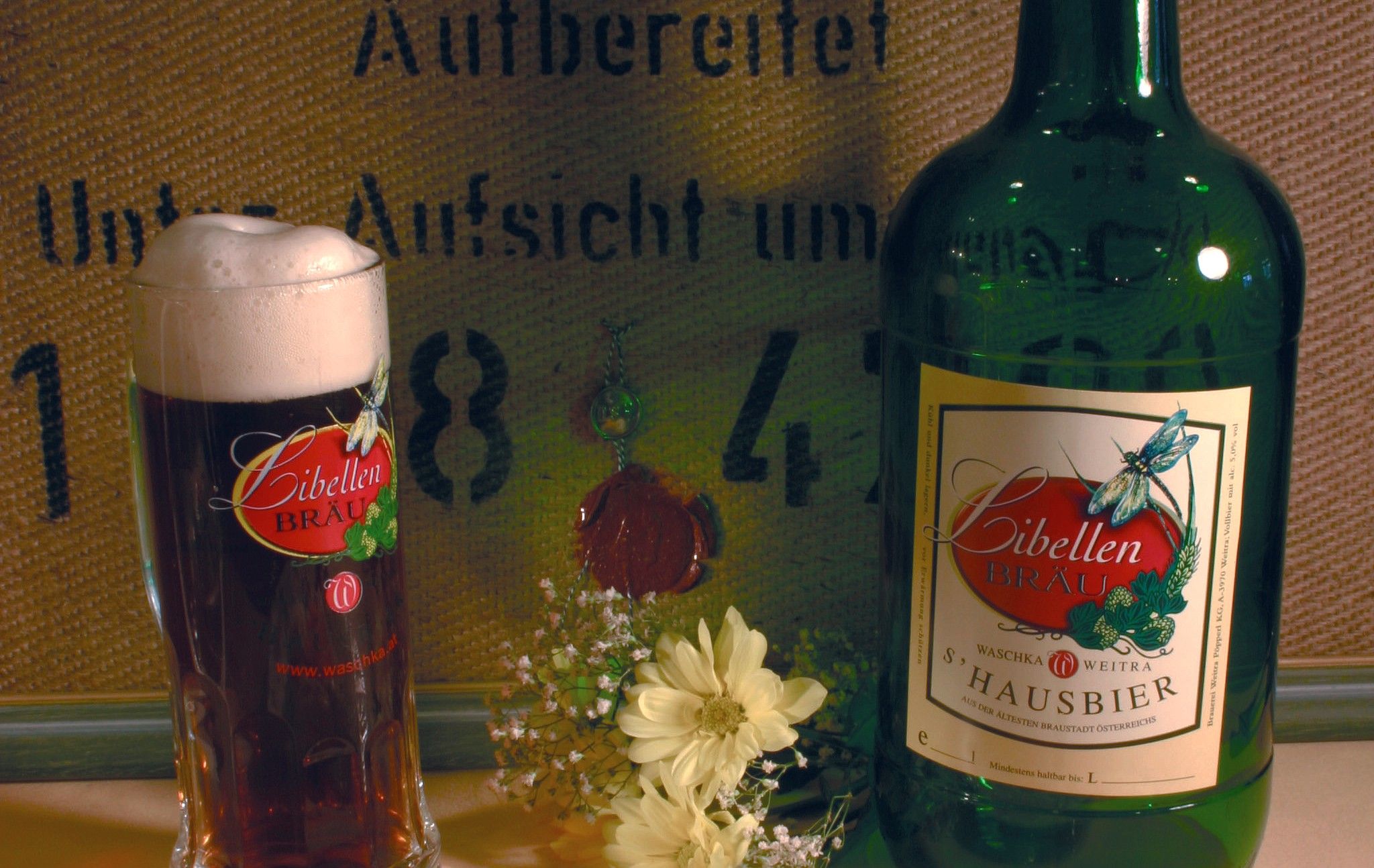 Ein Glas und eine Flasche Libellen Bräu Bier mit Blumen im Vordergrund.