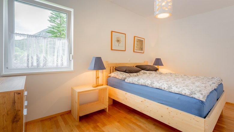 Ein gem&uuml;tliches Schlafzimmer mit einem Holzbett, blauer Bettw&auml;sche und zwei Nachttischlampen. Ein Fenster mit Blick auf B&auml;ume und Berge.