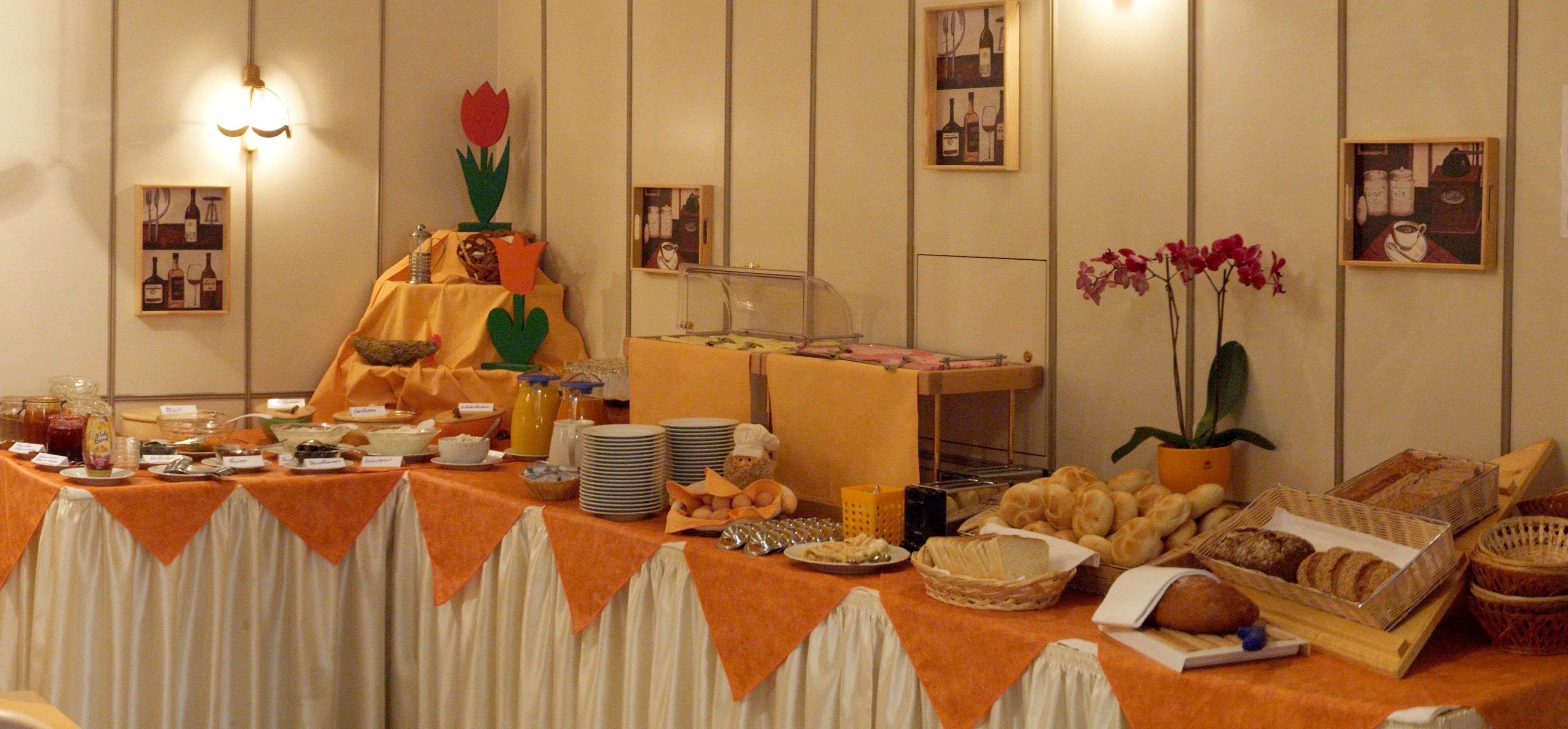 Ein reichhaltiges Frühstücksbuffet mit Brot, Brötchen, Aufstrichen und Getränken auf einem Tisch mit orangefarbenem Tuch.