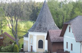 Eine kleine Kapelle mit spitzem Dach, umgeben von B&auml;umen und H&auml;usern.