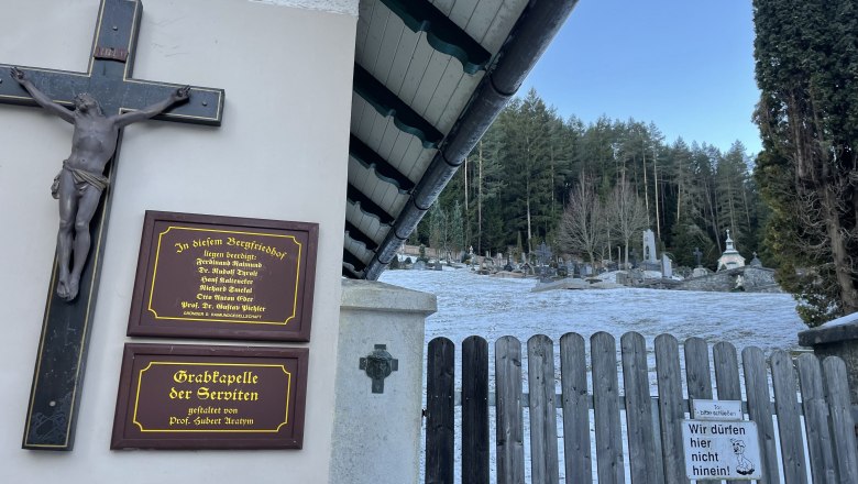 Eingang zum Bergfriedhof Gutenstein mit Holztor, Kreuz und Gedenktafeln. Im Hintergrund sind Gräber und Bäume zu sehen.