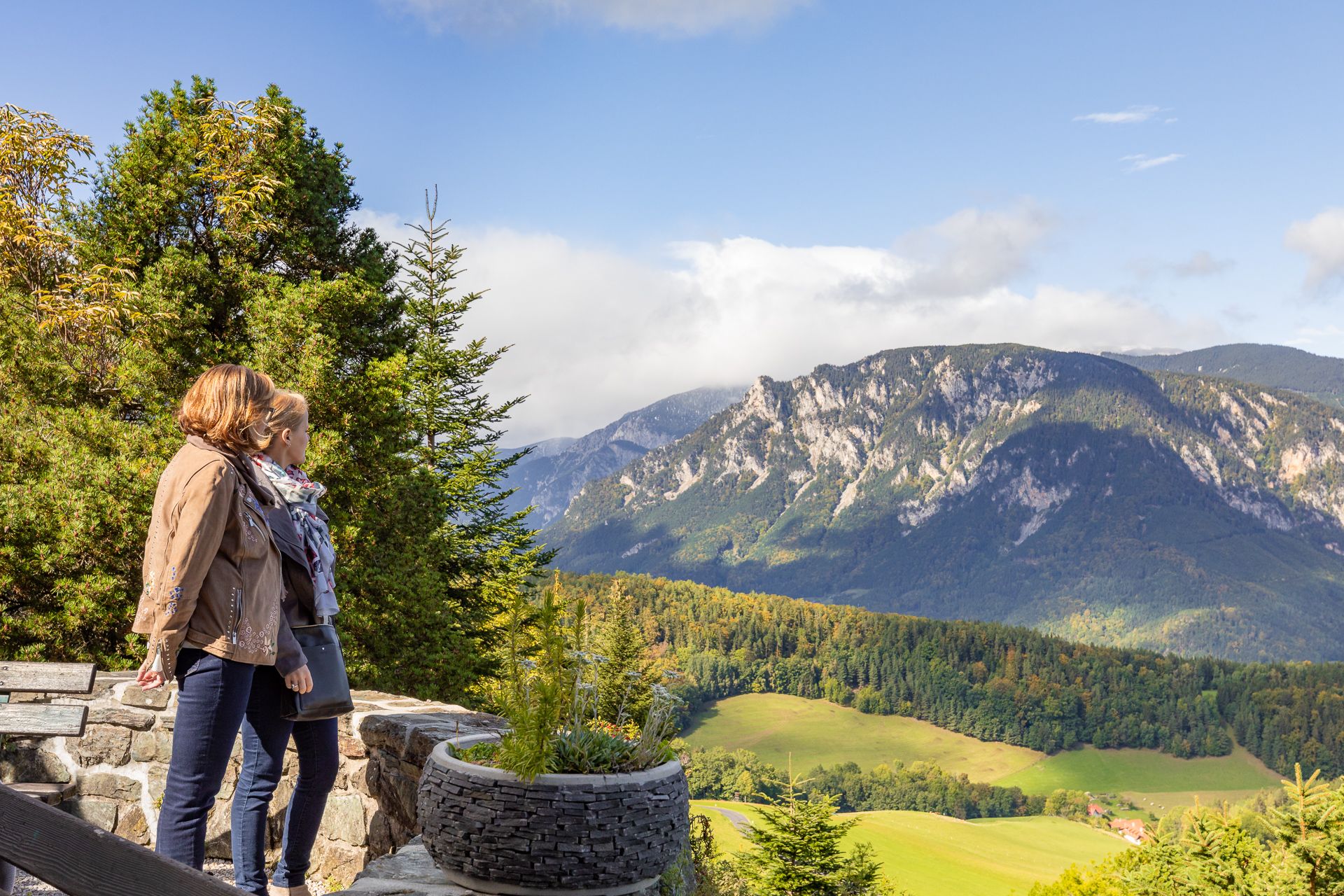 Zwei Personen stehen auf einer Terrasse und blicken auf eine Berglandschaft mit Wäldern und Wiesen.
