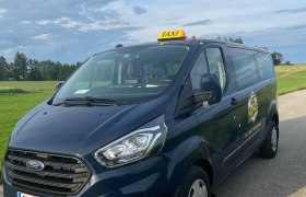 Ein blaues Ford-Taxi steht auf einer Landstra&szlig;e vor einer gr&uuml;nen Wiese und einem bew&ouml;lkten Himmel.