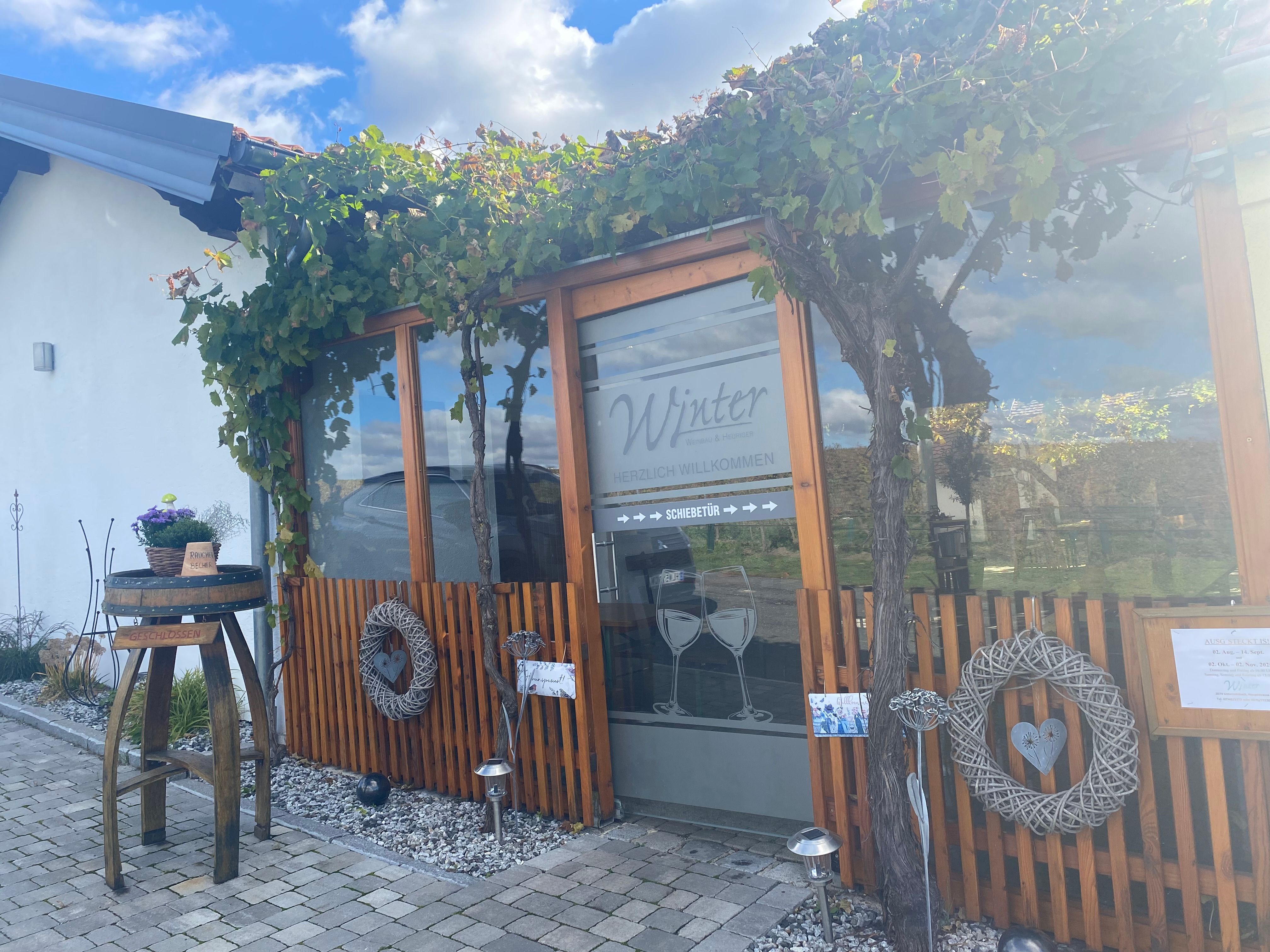 Eingang eines Gebäudes mit Holzverkleidung und Weinreben, dekoriert mit Kränzen und einem Tisch mit Blumen.