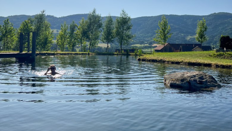 Person schwimmt in einem Teich mit Bäumen und Hügeln im Hintergrund.