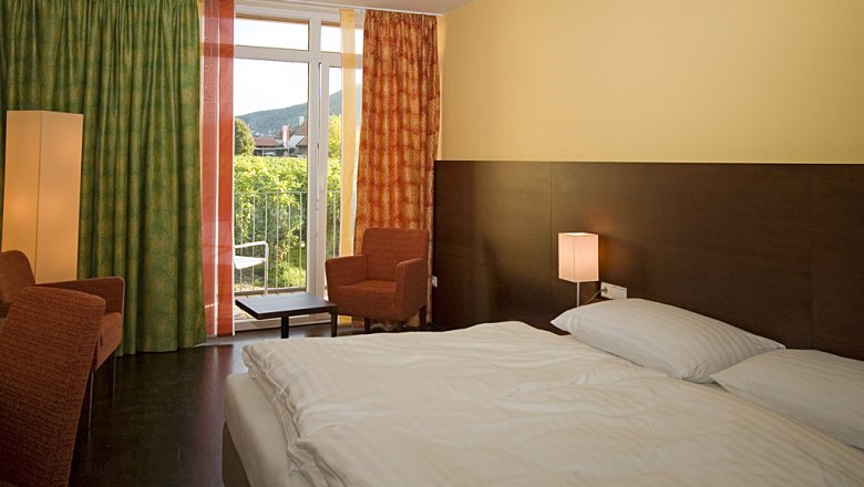 Hotelzimmer mit Doppelbett, zwei Sesseln, Vorhängen und Balkon mit Ausblick auf grüne Landschaft.