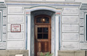 Eingangstür eines verzierten Gebäudes mit Holztür und dekorativen Malereien, umgeben von einem Holzzaun und Farnen.