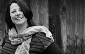 Schwarz-wei&szlig;-Foto einer l&auml;chelnden Frau mit Schal vor Holzwand.