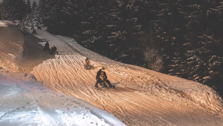 Menschen rodeln nachts auf einer beleuchteten Schneepiste am Semmering.