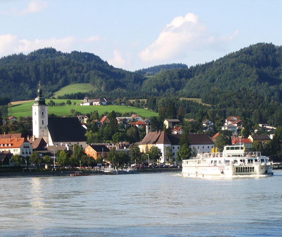 Stadt Grein mit Kirche und Schiff auf der Donau, umgeben von Hügeln.