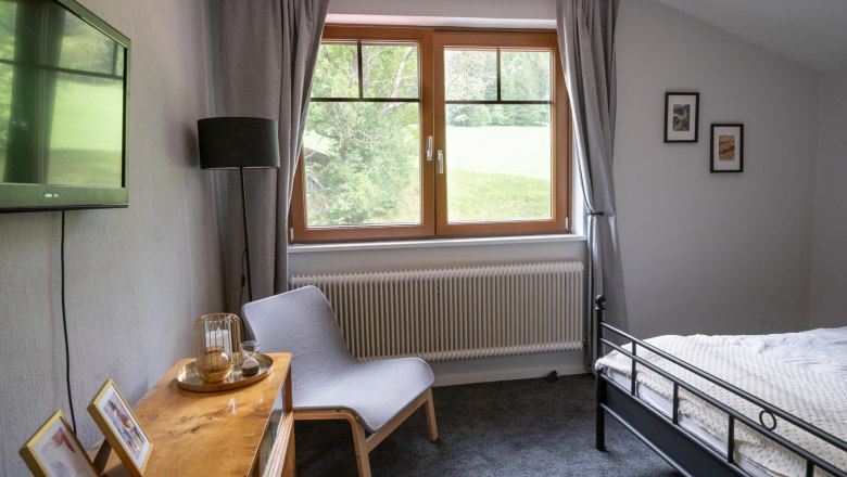 Schlafzimmer mit grauem Teppich, Bett, Sessel, Fernseher und Fenster mit Blick ins Grüne.
