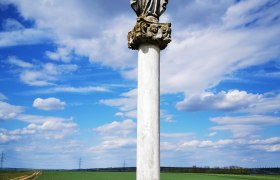 Eine Statue auf einer Säule in einer ländlichen Landschaft unter blauem Himmel mit Wolken.