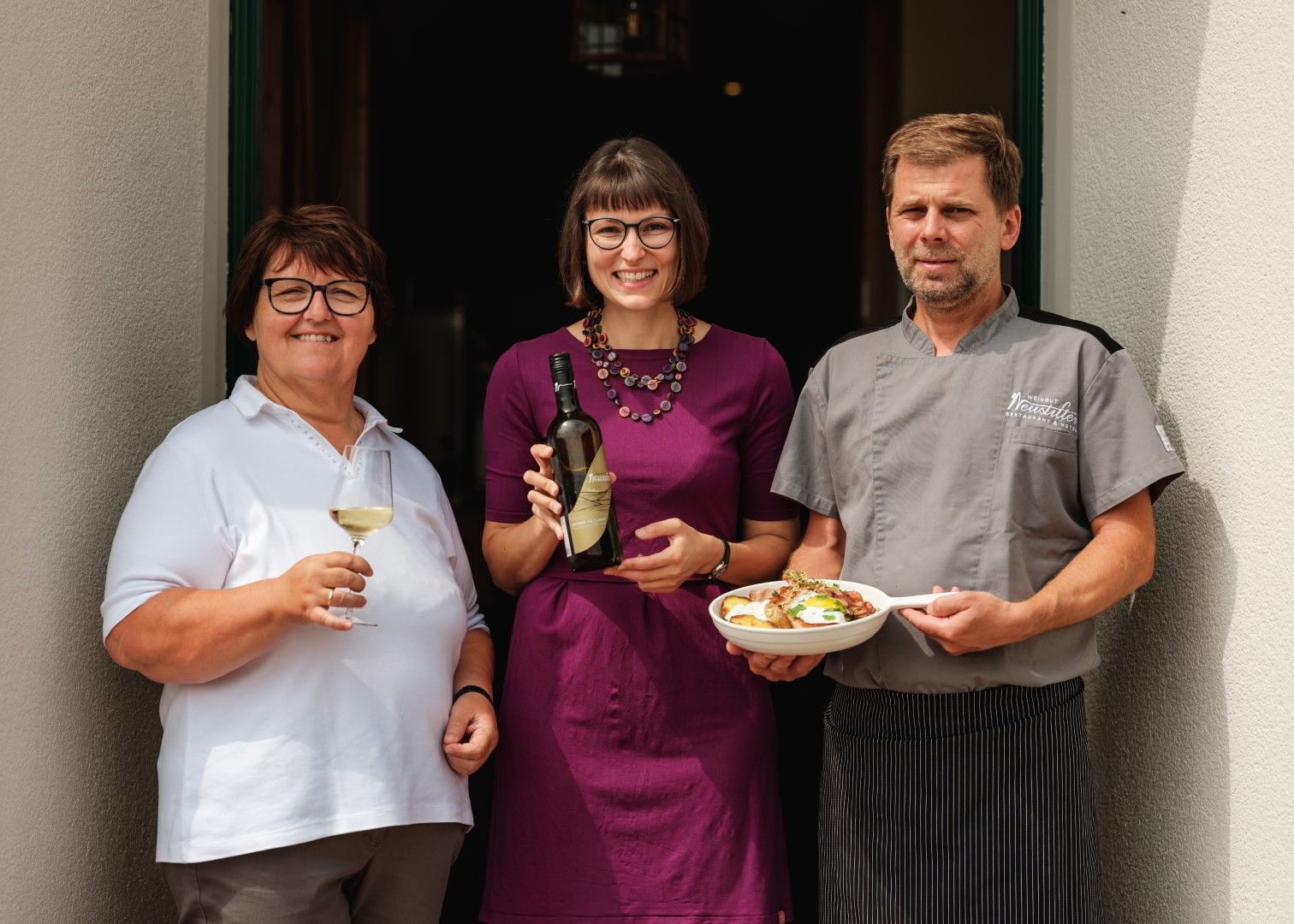 Drei Personen stehen vor einer Tür. Eine Frau hält ein Glas Wein, eine andere eine Weinflasche, und ein Mann trägt einen Teller mit Essen.