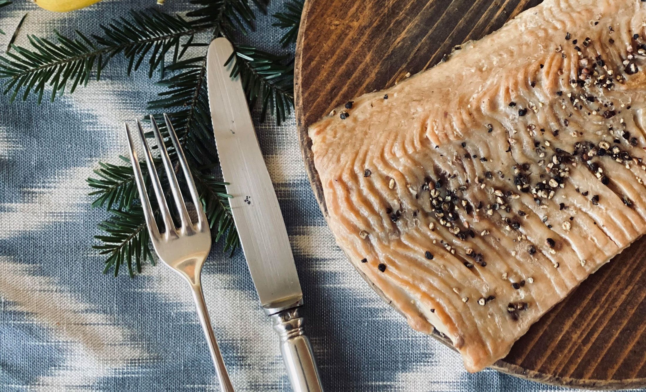 Nahaufnahme eines geräucherten Karpfenfilets auf einem Holzteller, dekoriert mit Pfefferkörnern. Daneben liegen eine Gabel und ein Messer auf einem blauen Tuch.