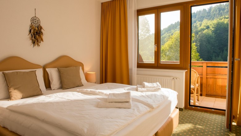Ein gem&uuml;tliches Doppelzimmer mit zwei Betten, einem Balkon mit Blick auf bewaldete H&uuml;gel und einem Traumf&auml;nger an der Wand.