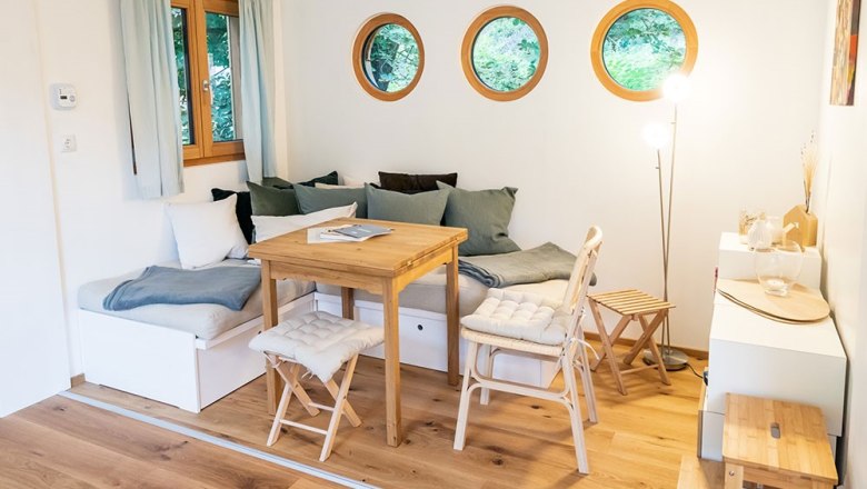 Gem&uuml;tlicher Sitzbereich mit Holztisch, St&uuml;hlen und runden Fenstern.