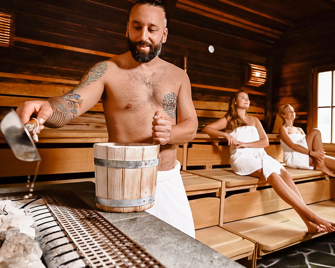 Mann gießt Wasser in Sauna, zwei Frauen sitzen entspannt.