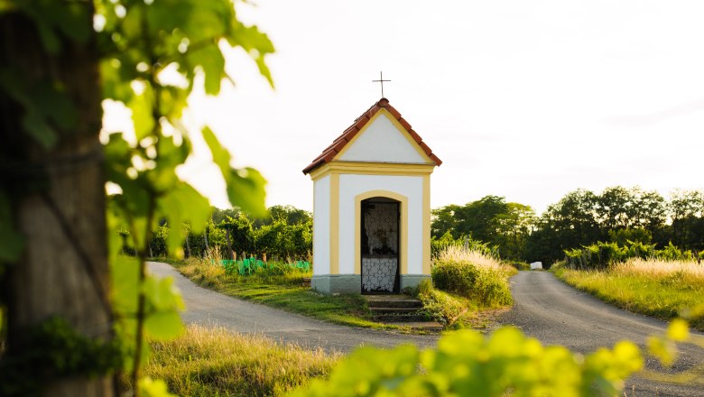 Kleine Kapelle an einem Weg in einer l&auml;ndlichen Landschaft, umgeben von gr&uuml;nen B&auml;umen und Pflanzen.