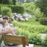 Ein Paar sitzt auf einer Holzbank in einem gr&uuml;nen Garten mit B&uuml;schen und B&auml;umen.