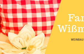 Rotes Kissen mit Karomuster und Handtuch in Blumenform auf einem Bett. Text: Familie Wißmann, Weinbau & Privatzimmer.