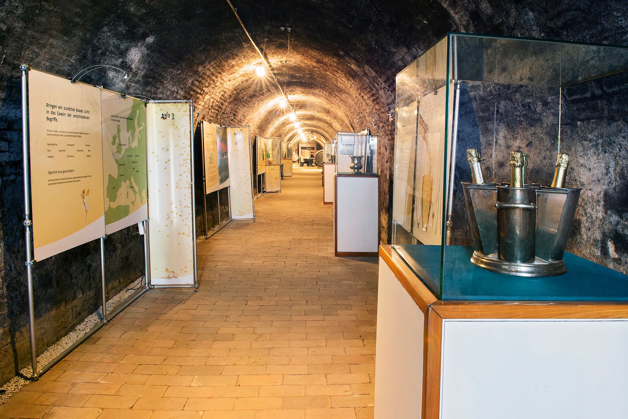 Ein Ausstellungsraum in einem Weinkeller mit Informationstafeln und Sektflaschen in Kübeln.