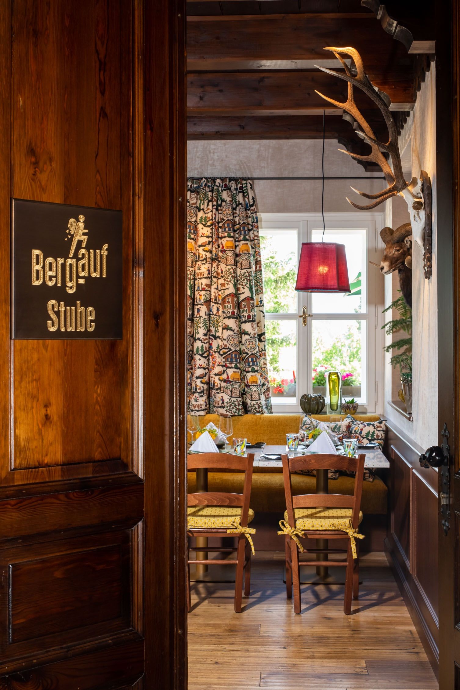 Ein gemütlicher Raum in einem Restaurant mit Holzmöbeln, einem Tisch mit gedecktem Geschirr, bunten Vorhängen und einem Geweih an der Wand.