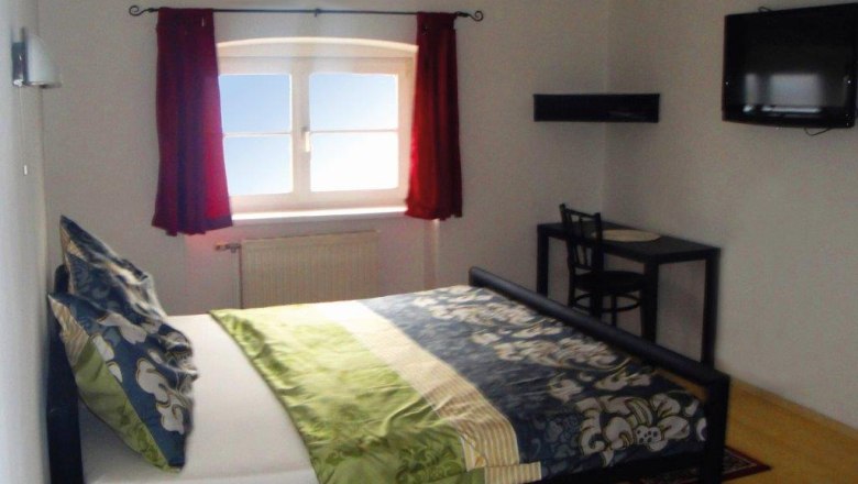 Ein Schlafzimmer mit einem Bett, einem Fenster mit roten Vorh&auml;ngen, einem kleinen Tisch mit Stuhl und einem an der Wand montierten Fernseher.