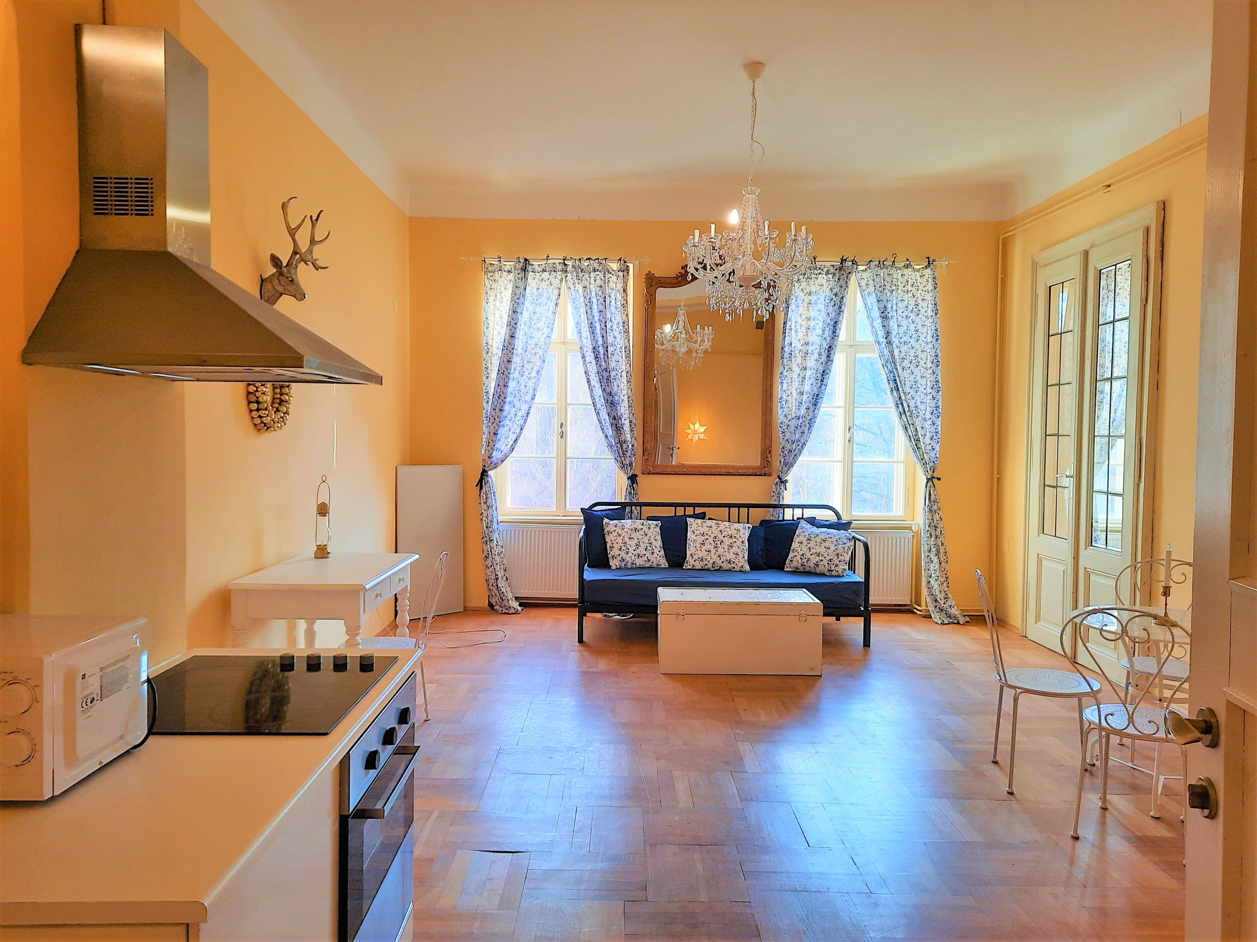 Wohnzimmer mit Küche, Esstisch und Sofa, gelbe Wände, Kronleuchter, Fenster mit Vorhängen.