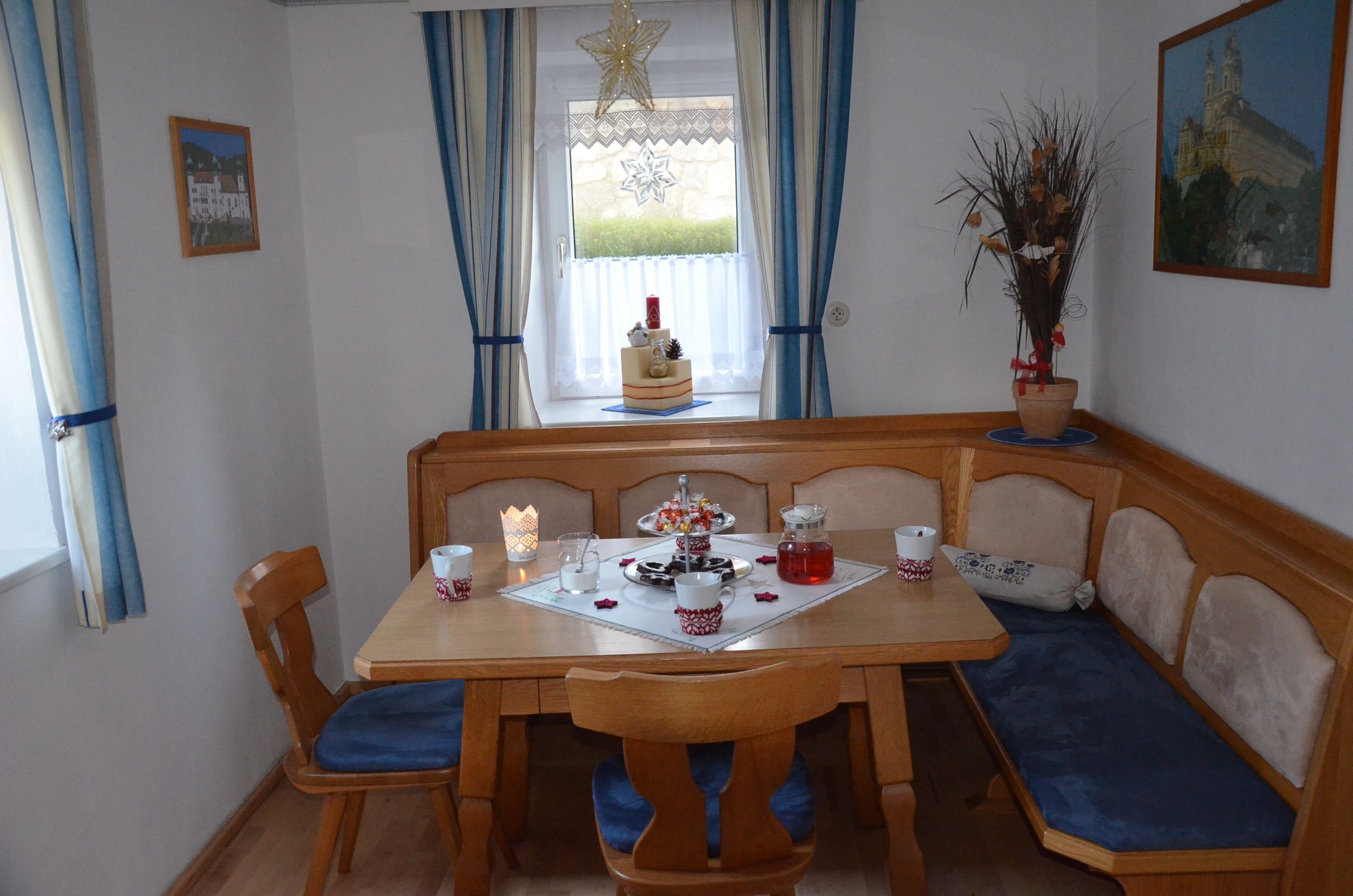 Gemütliches Esszimmer mit Holztisch, Eckbank und blauen Polstern. Fenster mit blauen Vorhängen und weihnachtlicher Dekoration.