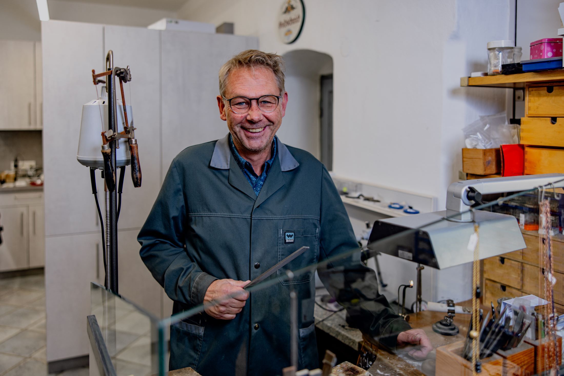 Ein Mann in einer Werkstatt mit Brille und Arbeitskittel, lächelnd, umgeben von Werkzeugen und Schmuck.