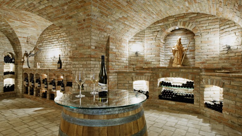 Ein Gew&ouml;lbekeller mit Ziegelw&auml;nden, Weinflaschen in Regalen und einem Fass mit Glasplatte als Tisch.