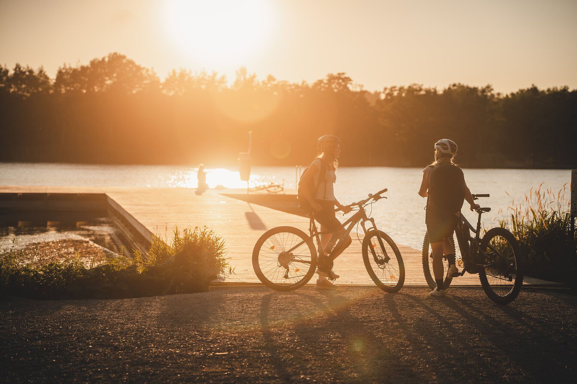 Zwei Radfahrerinnen mit Helmen stehen mit ihren Rädern bei tief stehender Sonne am Ufer des Viehofner Sees in St. Pölten.
