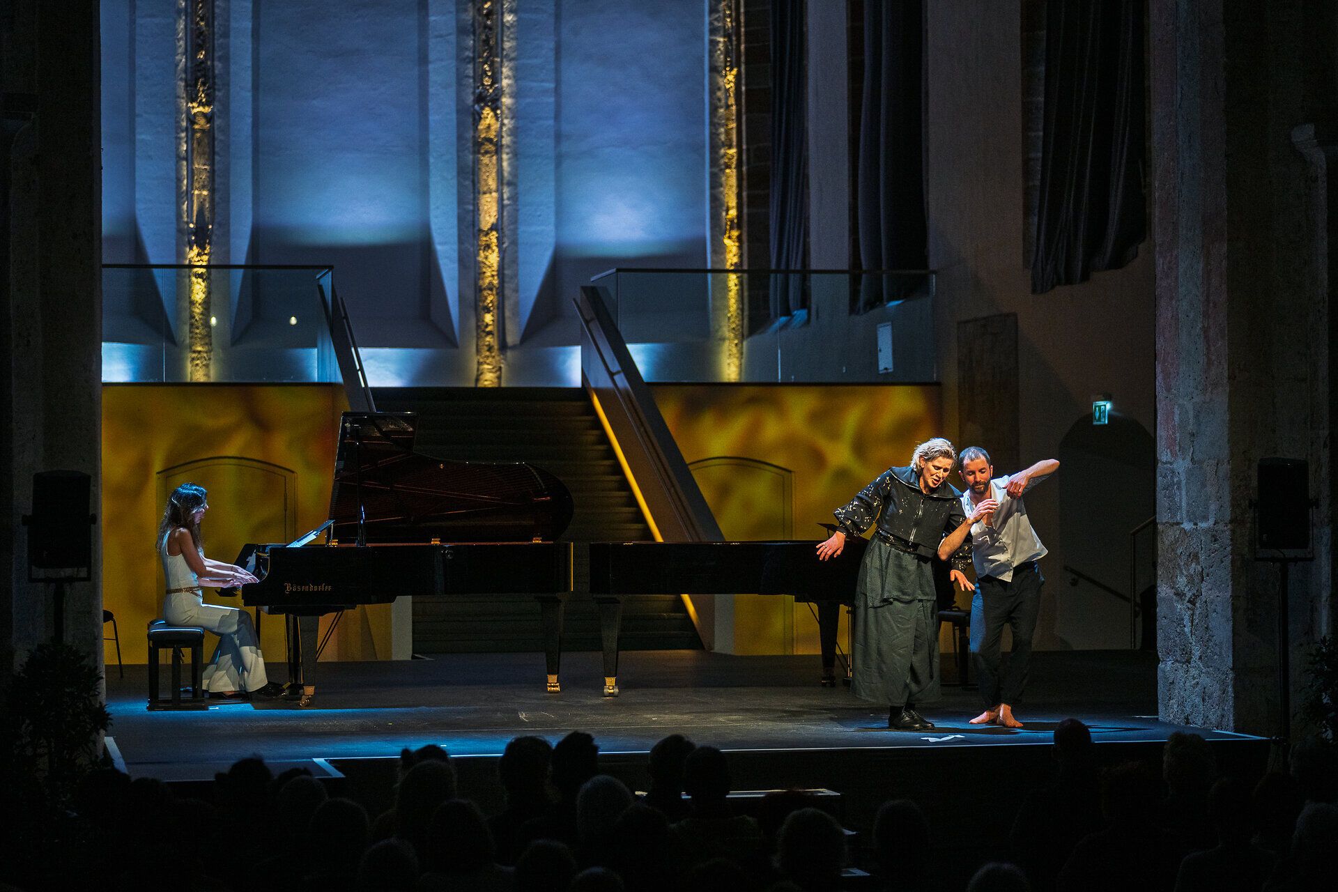 Szenische Musiktheateraufführung beim Imago Dei Festival: Pianistin am Flügel, Sängerin und Tänzer auf einer beleuchteten Bühne vor Publikum.