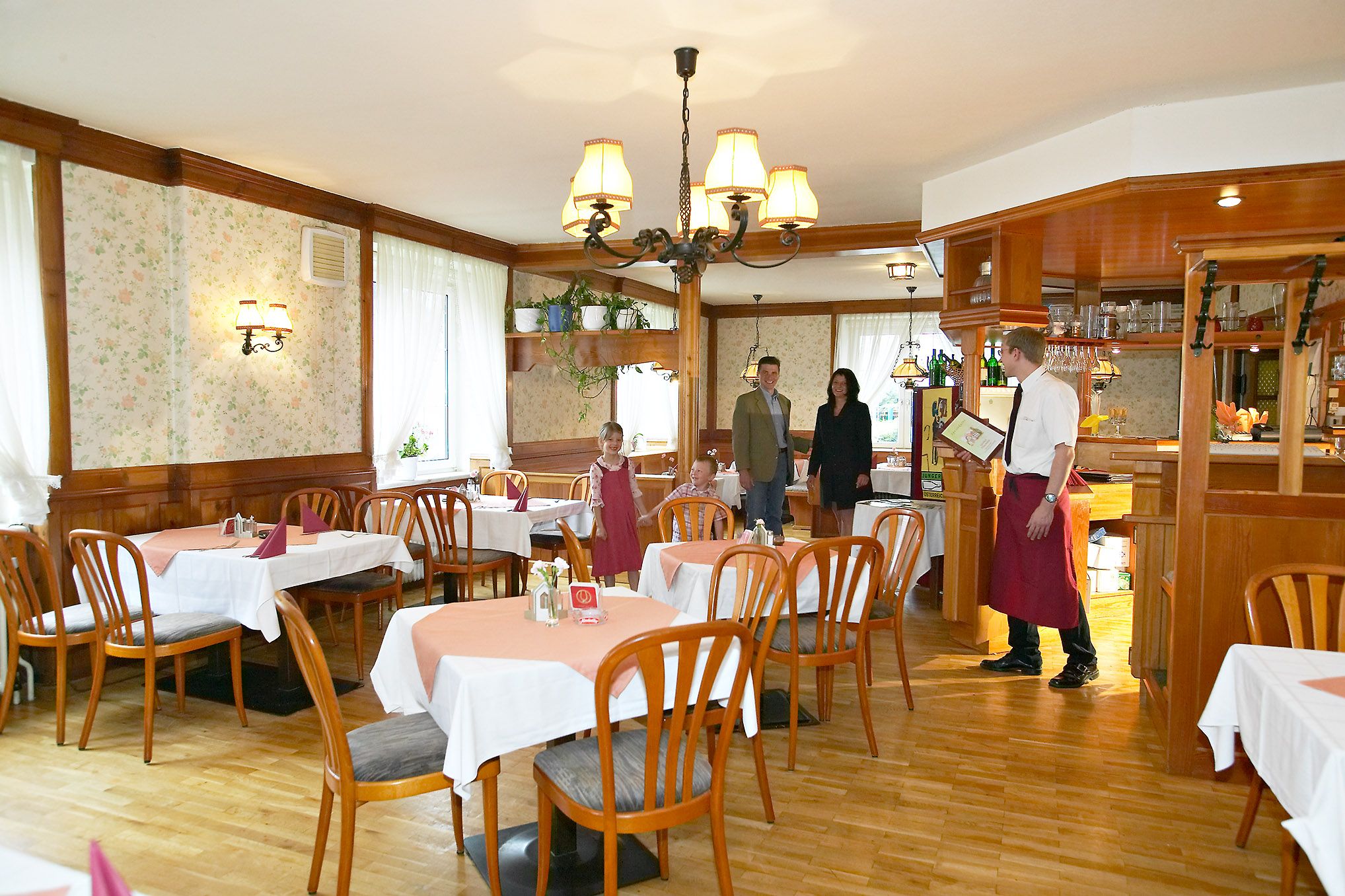 Ein rustikales Restaurant mit Holzmöbeln und Tischdecken. Ein Kellner begrüßt eine Familie am Eingang.