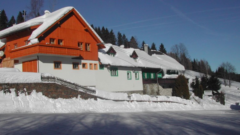Ein verschneites Gasthaus mit rotem Holzgiebel und weißen Wänden am Feistritzsattel, umgeben von Bäumen und blauem Himmel.