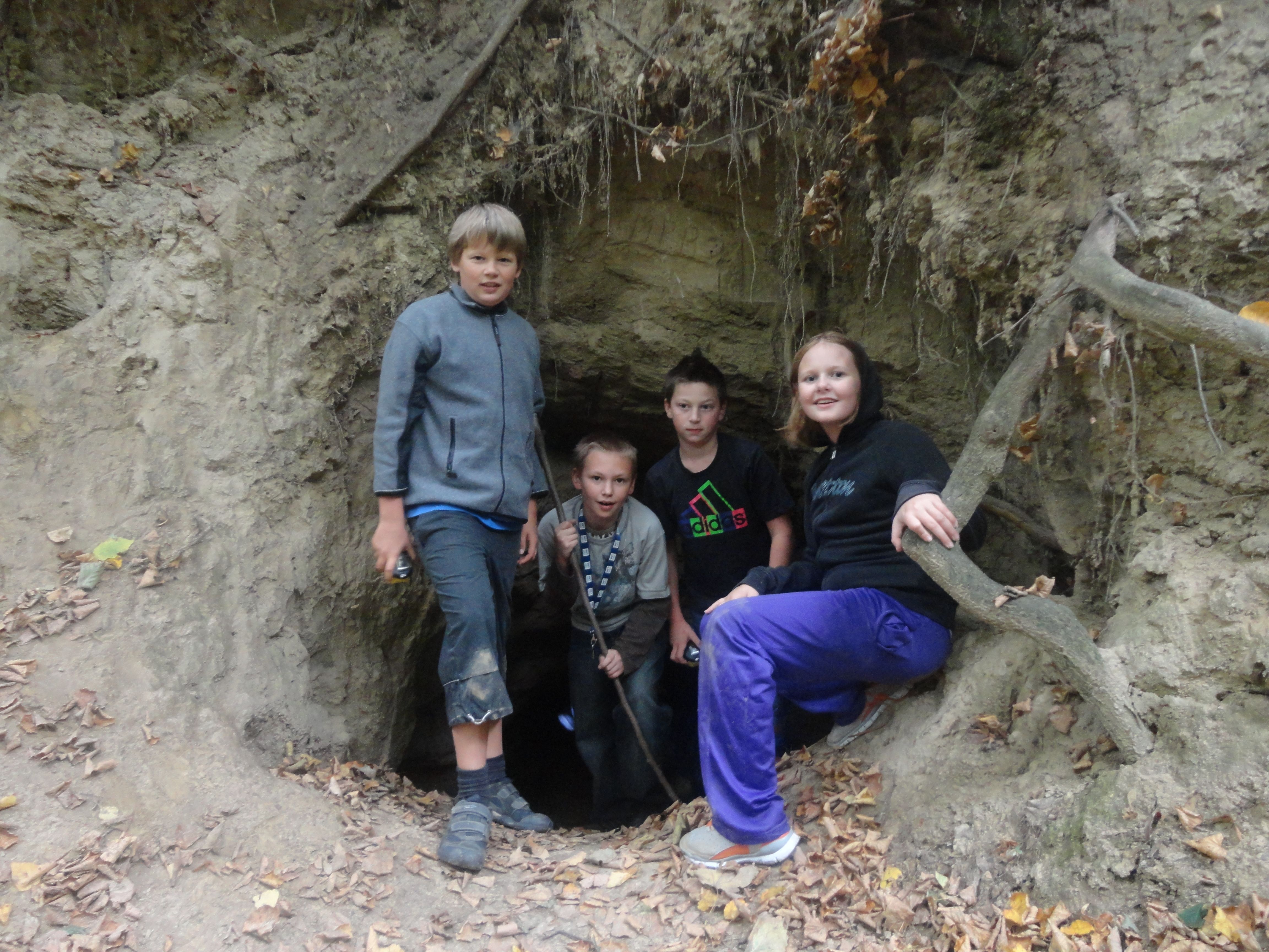 Vier Kinder stehen am Eingang einer kleinen Höhle im Wald.