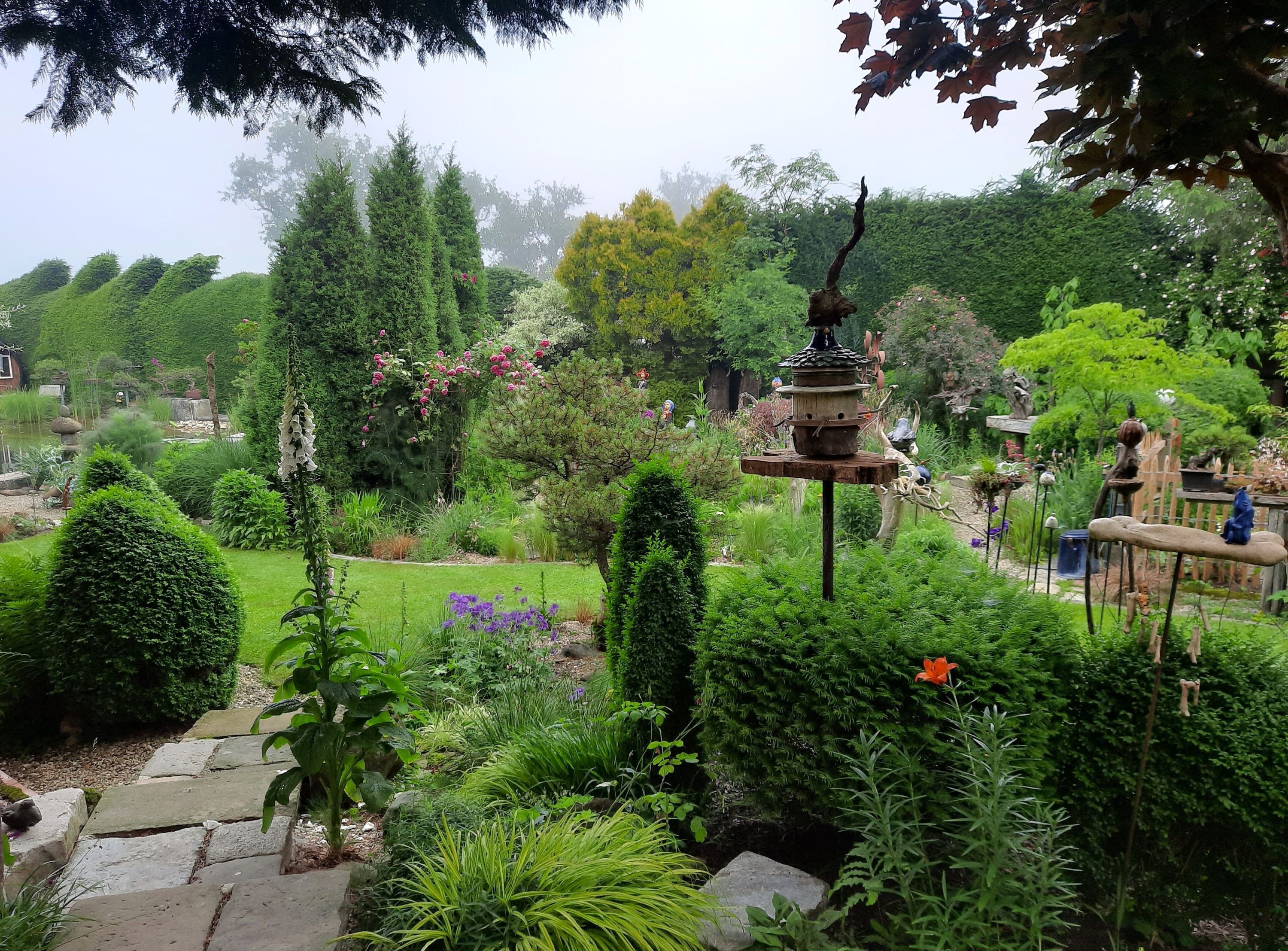 Ein üppiger Garten mit verschiedenen Pflanzen, Bäumen und Dekorationen. Ein Vogelhaus steht im Vordergrund.