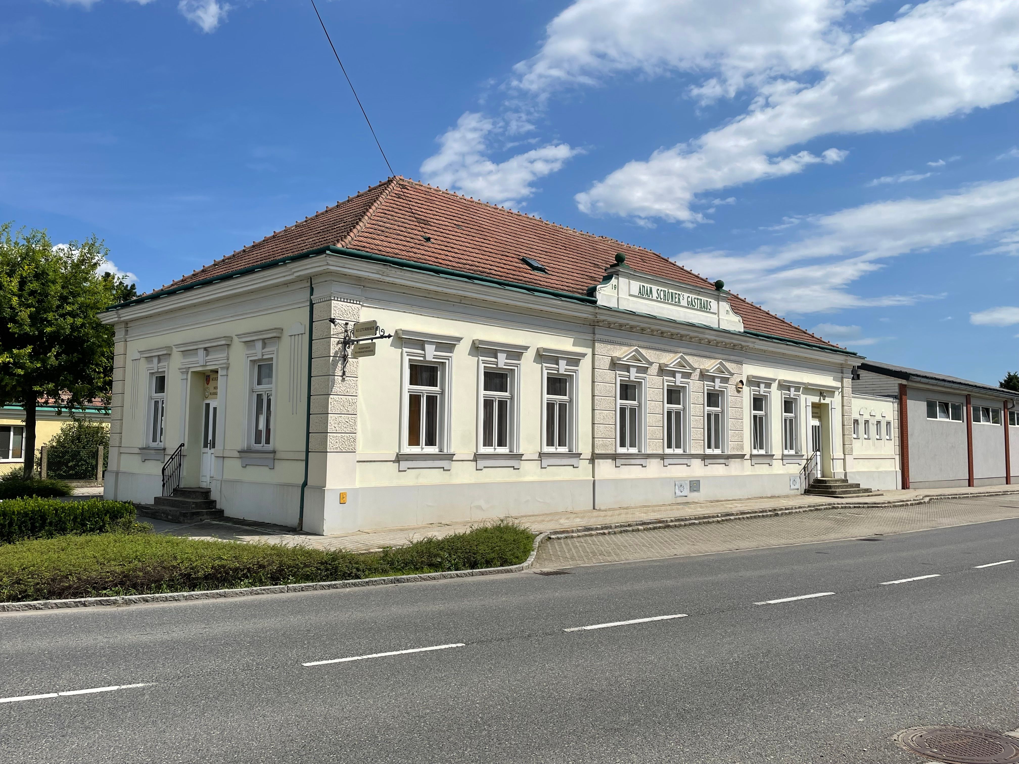 Das Kulturhaus, ein helles historisches Gebäude mit rotem Ziegeldach, steht an einer Straße unter blauem Himmel.
