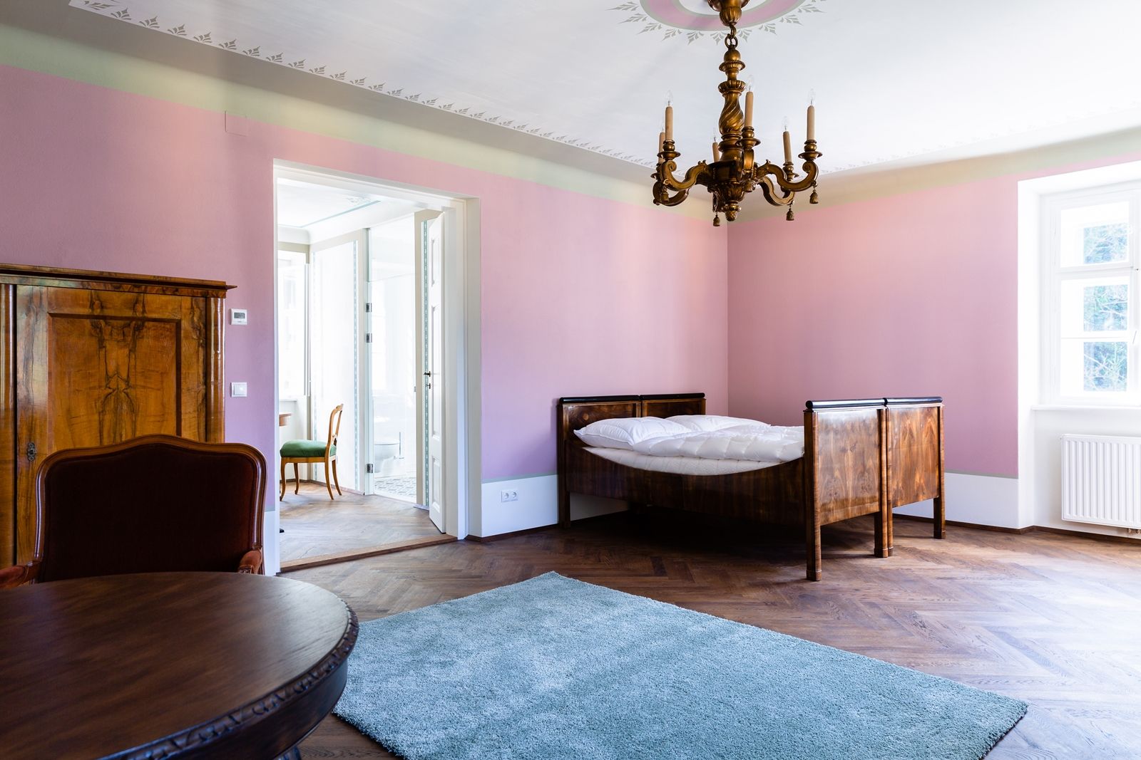 Ein elegantes Zimmer mit rosa Wänden, einem Holzbett, einem Kronleuchter und einem Teppich auf Holzboden.