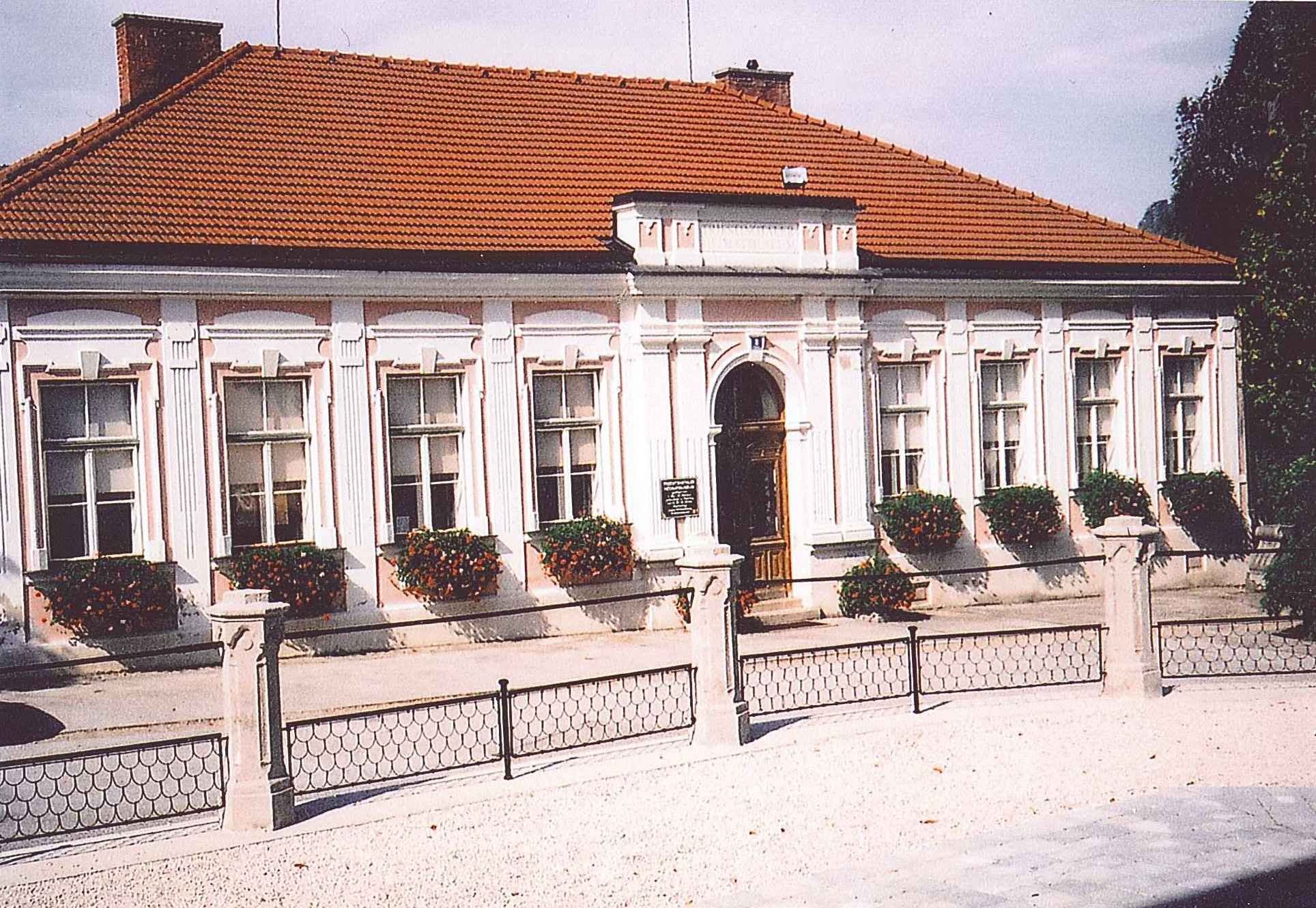 Historisches Gebäude mit rotem Ziegeldach und weißen Fensterrahmen, umgeben von Blumen.