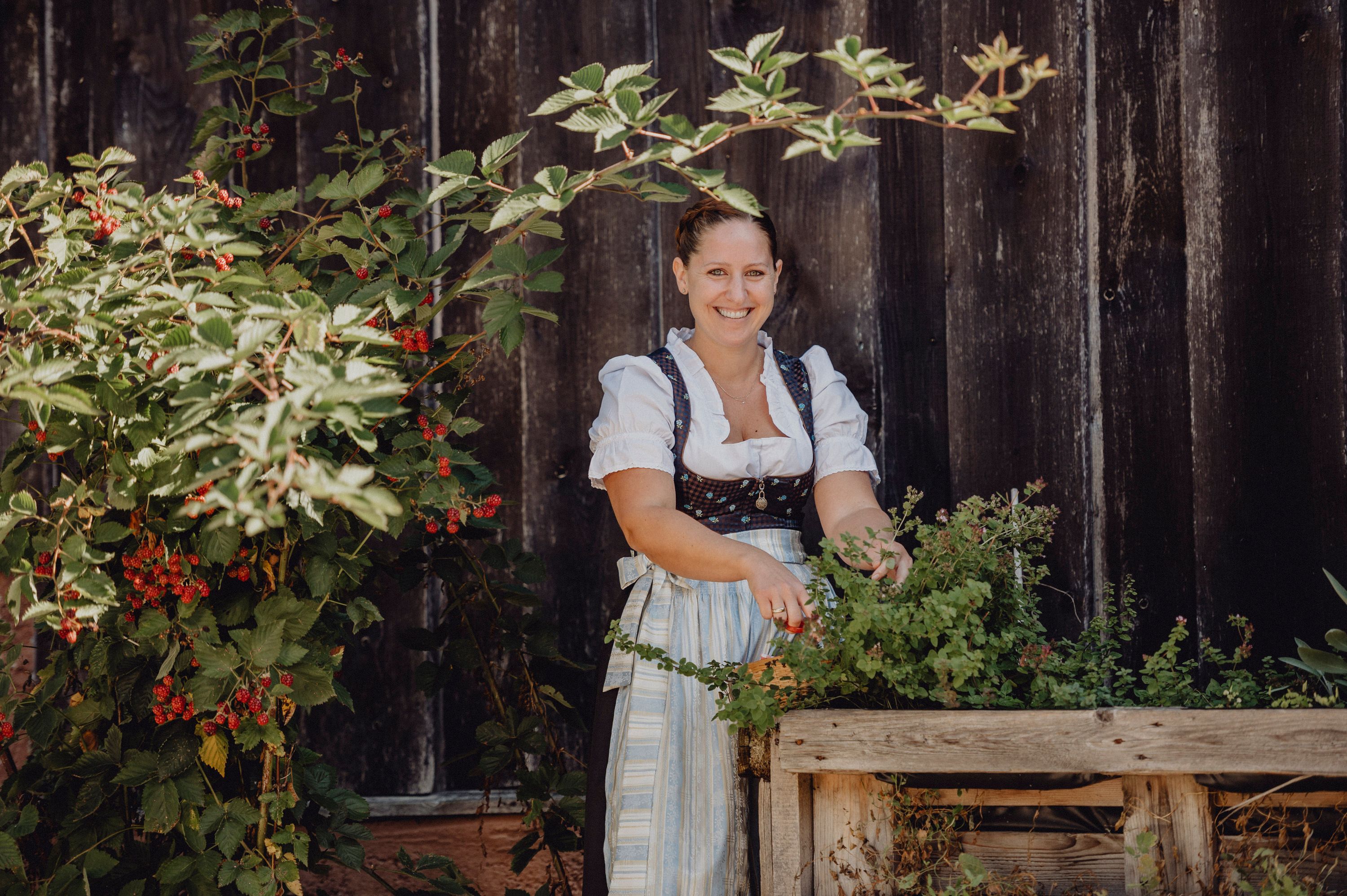 Frau in traditioneller Kleidung steht lächelnd neben einem Kräuterbeet vor einer Holzwand.