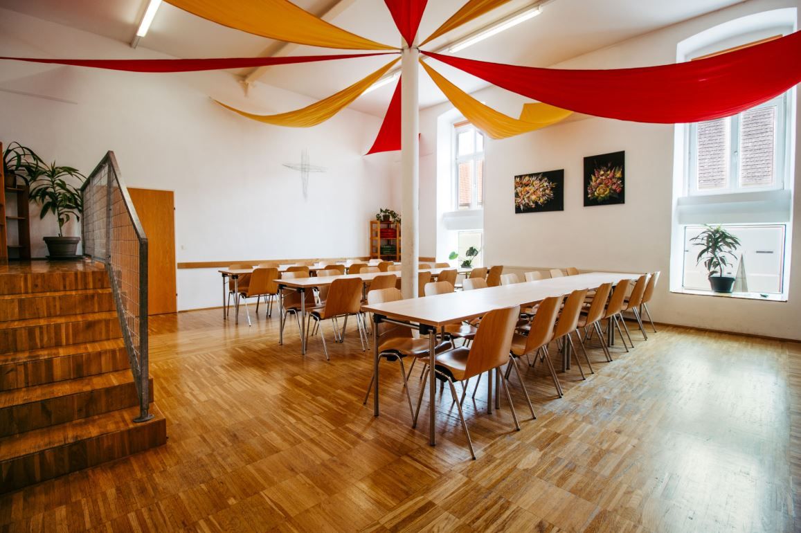 Ein heller Speisesaal mit Holzboden, Tischen und Stühlen. An der Decke hängen rote und gelbe Stoffbahnen.