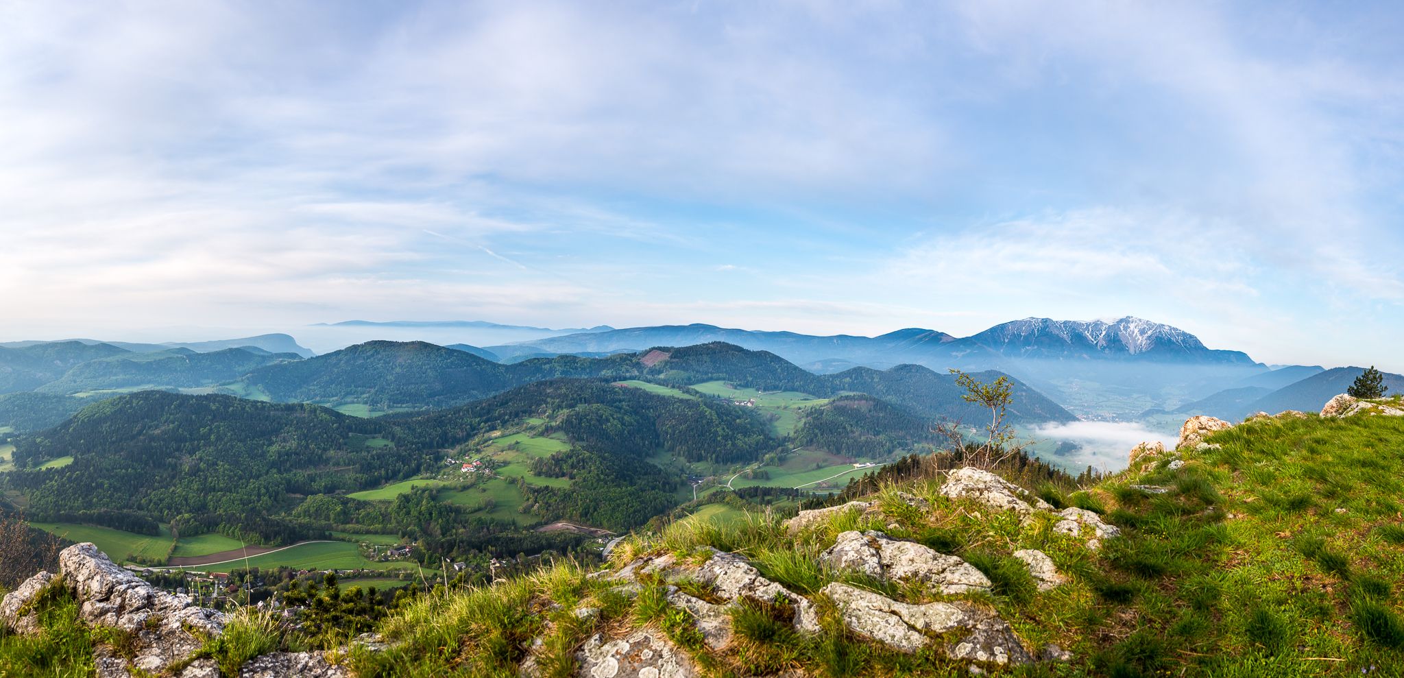 Panoramablick auf eine grüne Berglandschaft mit bewaldeten Hügeln und einem schneebedeckten Berg im Hintergrund unter blauem Himmel.